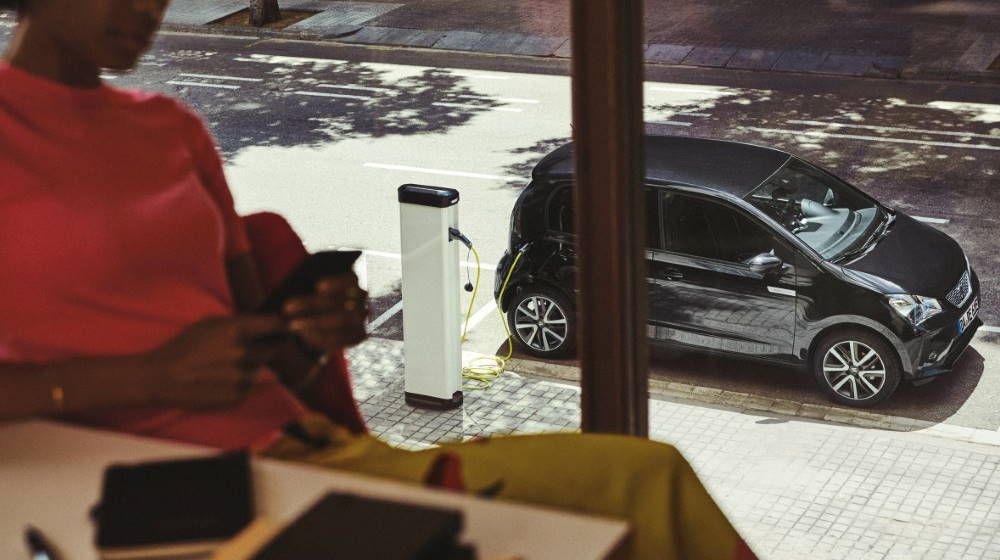 Frau sitzt im Cafe während draußen ihr schwarzer SEAT Mii electric an einer Ladesäule aufgeladen wird