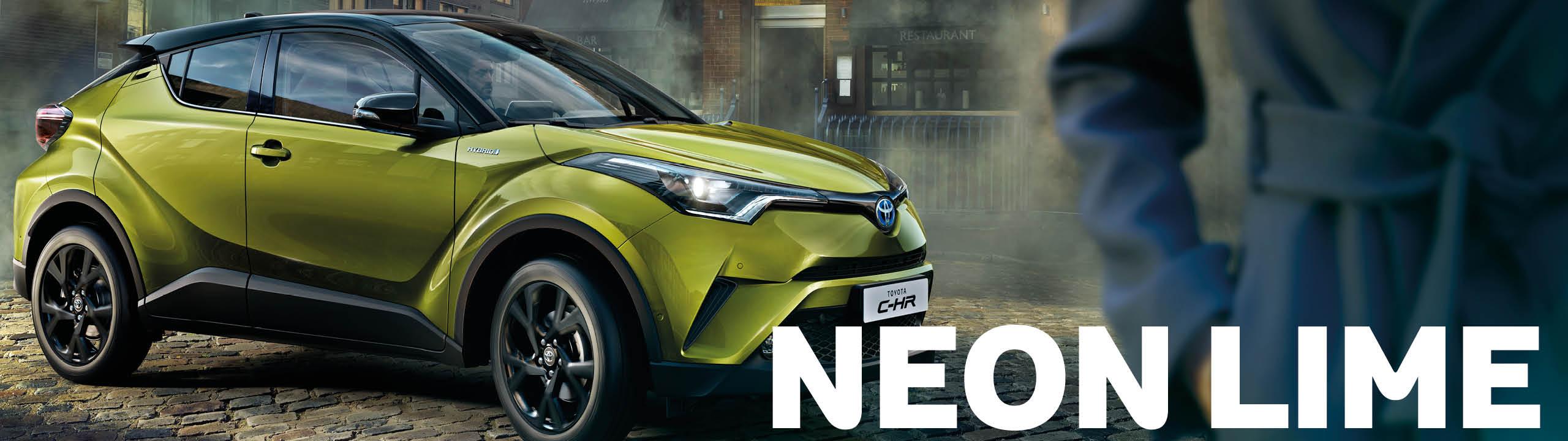 Toyota C-HR Neon Lime, grün mit schwarzem Dach, steht in der Seitenansicht auf Pflasterstein, in der rechten Bildschirmhälfte ist ein Teil einer Person in einem Mantel zu sehen